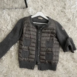 Grå cardigan jacka från Moncler - Säljer en stilren grå cardigan jacka från Moncler med quiltad framsida och stickade ärmar. Jackan har knappar framtill och två fickor. Perfekt för kyliga dagar!
