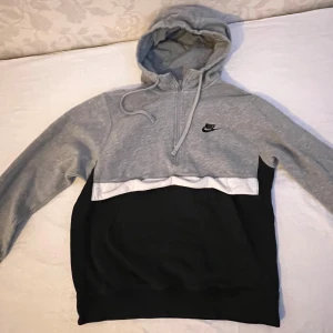 Grå och svart hoodie från Nike - Säljer en snygg grå och svart hoodie från Nike med dragkedja och huva. Tröjan har en vit rand över bröstet och en liten Nike-logga på framsidan. Perfekt för en avslappnad stil.