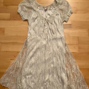 Spetsklänning - Charmig klänning med spetsdetaljer och knappar framtill. Den har korta ärmar och ett knytband i midjan. Perfekt för somriga dagar.
