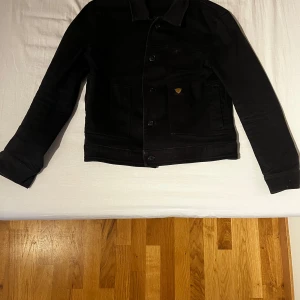 Svart jeansjacka från Tiger of Sweden - Snygg svart jeansjacka från Tiger of Sweden med klassisk design. Jackan har knappar framtill och en diskret logga i gult på bröstfickan. Perfekt för en stilren look.