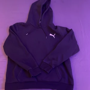 Svart hoodie från Puma - Säljer en klassisk svart hoodie från Puma med logotyp på bröstet. Tröjan har en bekväm passform med långa ärmar och en praktisk känguruficka framtill. Perfekt för en avslappnad stil.