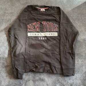 Svart sweatshirt från Tommy Jeans - Säljer en svart sweatshirt från Tommy Jeans med texten 'New York' i rött och '1985' i vitt. Tröjan har långa ärmar och en rund halsringning. Perfekt för en avslappnad stil. Stor till passformen, passar bättre på M än S