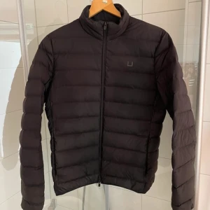 Svart pufferjacka från UBR - Snygg svart pufferjacka/dunjacka från UBR med dragkedja framtill. Jackan har en quiltad design och långa ärmar, perfekt för höstliga och vår dagar. Den är stilren och lätt att matcha med olika outfits.