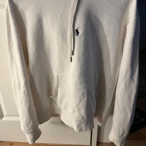 Vit hoodie från Polo Ralph Lauren - Säljer en stilren vit hoodie från Polo Ralph Lauren med det klassiska logotypbroderiet på bröstet. Tröjan har en känguruficka och justerbar huva med snören. Perfekt för en avslappnad look.