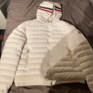 Vit dunjacka med huva - Snygg vit dunjacka från Moncler med huva och dragkedja. Jackan har ett quiltat mönster och ribbade detaljer. Perfekt för kyliga dagar med stil.