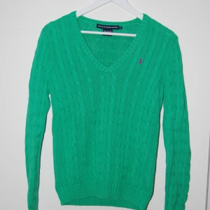 Grön stickad tröja från Ralph Lauren Sport - Säljer en snygg grön stickad tröja från Ralph Lauren med en lila broderad logga på bröstet. Tröjan har en v-ringad design och långa ärmar, perfekt för en stilren look. Den är i mycket bra skick och gjord i ett väldigt mjukt och bekvämt material. Den är i storlek S