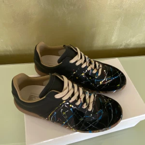 Svarta sneakers från Maison Margiela - Unika svarta sneakers från Maison Margiela med färgglada stänk i blått, gult och rött. Skorna har en klassisk snörning och en bekväm gummisula. Perfekta för dig som vill sticka ut med en konstnärlig touch. De är använda runt 3-5 gånger men inga skador priset är inte fast kan alltid diskuteras. Var inte rädd att ställa frågor.💪😃