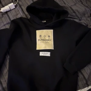 Burberry hoodie - Säljer en stilren svart hoodie från Burberry med en stor beige logga på framsidan. Hoodien har en klassisk design med långa ärmar och en bekväm huva. Perfekt för en avslappnad och trendig look.