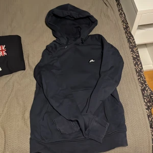 Mörkblå hoodie från J.Lindeberg - Säljer en stilren mörkblå hoodie från J.Lindeberg. Tröjan har en klassisk design med en liten logotyp på bröstet och en bekväm huva. Perfekt för en avslappnad look. 