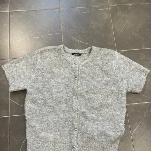 Grå kofta från Gina Tricot - Mysig grå kofta från Gina Tricot med korta ärmar och knappar framtill. Perfekt för lager-på-lager under kyligare dagar. Enkel och stilren design som passar till det mesta.