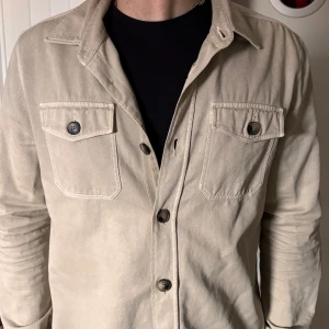 Beige overshirt från Morris - Säljer en stilren beige overshirt från Morris. Jackan har långa ärmar och knappar framtill samt två bröstfickor med knappar. Perfekt för en avslappnad look. Ord pris 2000sek