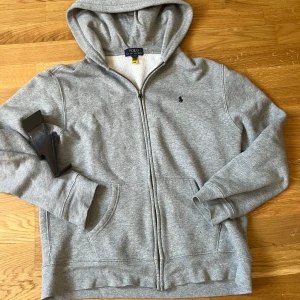 Grå hoodie från Polo Ralph Lauren - Säljer en stilren grå hoodie från Polo Ralph Lauren med dragkedja och huva. Tröjan har en mjuk insida och två praktiska fickor framtill. Perfekt för en avslappnad look.