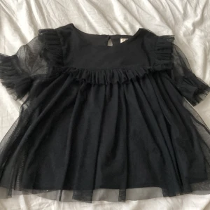 Svart volangblus med tyllärmar - Säljer en svart blus med volangdetaljer och transparenta tyllärmar. Blusen har rund halsringning och en liten öppning med knapp i nacken. Perfekt för dig som gillar en luftig och söt stil.