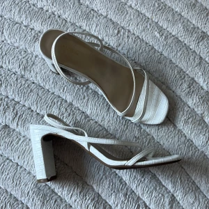Vita sandaler med klack  - Säljer ett par vita klackskor. Skorna har tunna remmar och ett stilrent krokodilpräglat mönster. Perfekta för dig som gillar minimalistisk och trendig design. Använda men bra skick. Ca 8cm klack. 