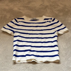 Vit och blå randig ribbad topp från Zara - Säljer en vit ribbad topp från Zara med blå horisontella ränder. Toppen har korta ärmar och rund halsringning. Perfekt för en fräsch och avslappnad stil.