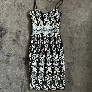Blommig spetsklänning från Lipsy London - Supersöt svart klänning från Lipsy London med tunna axelband och broderade blommor i vitt och ljusrosa. Klänningen har v-ringning och vacker spetsdetalj i midjan. Perfekt för dig som gillar romantisk stil och fina detaljer.