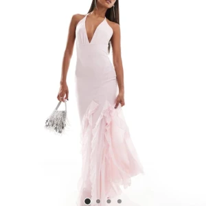 Ljusrosa långklänning med volangdetaljer - Säljer en elegant ljusrosa långklänning med djup v-ringning och smala axelband. Klänningen har volangdetaljer längs kjolen och en figurnära passform som framhäver siluetten. Perfekt för speciella tillfällen.