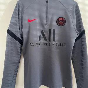Grå långärmad PSG fotbollströja från Nike - Säljer en grå långärmad Paris Saint-Germain fotbollströja från Nike med svart och rött PSG-emblem på bröstet. Tröjan har svarta detaljer på ärmarna och rött Nike märke. Tröjan har en halv dragkedja framtill.