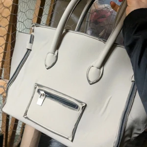 Beige handväska med dragkedja - Stilren beige handväska med två rejäla handtag och en praktisk ytterficka med dragkedja framtill. Väskan har svarta detaljer längs kanterna och är perfekt för att bära med sig det viktigaste till vardags.