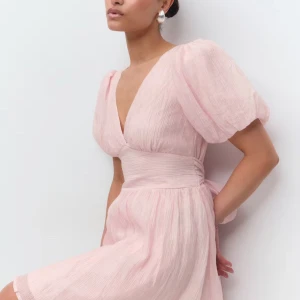 Helt slutsåld och oanvänd ljusrosa klänning från Ginatricot  - Säljer denna superfina ljusrosa klänning med v-ringning, puffiga kortärmar och omlottknytning i midjan från ginatricot. Klänningen har en öppen rygg med stor rosett och är perfekt inför midsommar! Aldrig använd och lapparna finns kvar, helt slutsåld på hemsidan!🩷