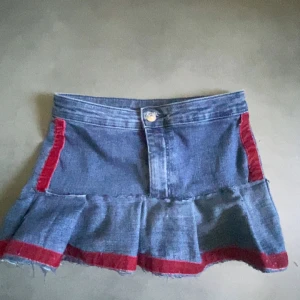 Jeanskjol med volang och röda detaljer - Säljer en kort jeanskjol med volang och råa kanter. Kjolen har röda band längs sidorna och nederkanten, samt klassisk knapp och fickor bak. Perfekt för dig som vill sticka ut med en unik denimlook.den är även gjord helt själv 