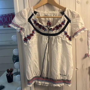 Vit blus med broderade blommor från Bondelid - Supersöt vit blus från Bondelid med broderade röda och blå blommor, svarta detaljer och knytband vid halsen. Blusen har korta ärmar och elastiska muddar vid ärmslut och nederkant. Perfekt för en somrig look!