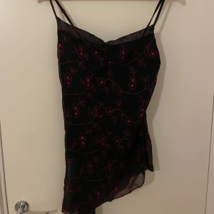 Svart asymmetrisk klänning med rosa blommor från Interco - Svart topp med tunna axelband och asymmetrisk nederkant. toppen har ett genomskinligt lager med broderade rosa blommor som ger en unik look. Perfekt för dig som vill sticka ut med en feminin och trendig stil.