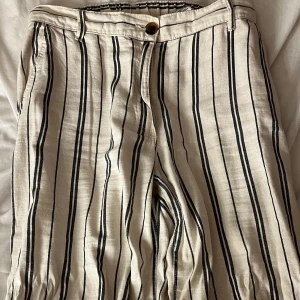Randiga linnebyxor med vida ben - Säljer ett par beige linnebyxor med svarta och grå vertikala ränder. Byxorna har normal passform, hög midja och vida ben. Stängs med knapp och dragkedja framtill och har fickor både fram och bak. Perfekta för varmare dagar.