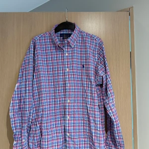 Rutig skjorta från Ralph Lauren - Säljer en klassisk rutig skjorta från Ralph Lauren i blått, rött och vitt. Skjortan har lång ärm, knappar framtill och broderad logga på bröstet. Perfekt för dig som gillar tidlös stil och vill ha något som sticker ut lite extra.