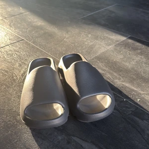 Yeezy slides - Säljer ett par gråa Adidas slides med öppen tå och räfflad sula. De är tillverkade i syntetmaterial och har en minimalistisk design med Adidas-loggan på fotbädden. Perfekta för sommaren och chill dagar.