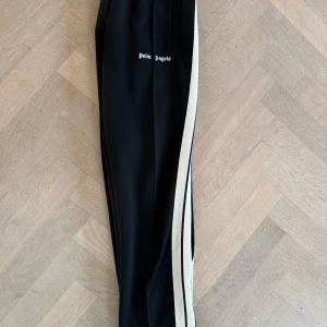 Svarta Palm Angels byxor med broderad logga - Palm Angels svarta byxor med beige rand längs sidorna och broderad logga på vänster ben. Byxorna har elastisk midja, raka ben och dragkedjor vid bensluten.  Nypris 5000 kr