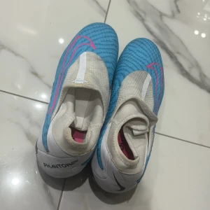 Nike Phantom GT2 Elite fotbollsskor blå/vit - Säljer ett par Nike Phantom GT2 Elite fotbollsskor i blått och vitt med rosa detaljer. Skorna har en strumpliknande passform utan traditionell snörning och en sula med fasta dobbar för spel på gräs. Perfekta för dig som vill ha kontroll och precision på planen.