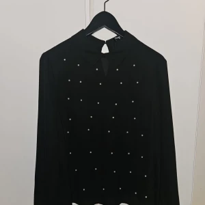 Svart blus med pärldetaljer från Fashion Union - Snygg svart blus från Fashion Union med långa ärmar och pärldetaljer framtill. Blusen har en liten öppning vid halsen och knäpps i nacken. Perfekt för dig som vill ha något stilrent med en twist.