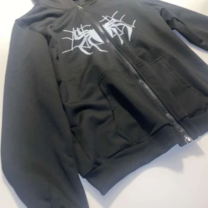 Svart hoodie med vitt spindelnätsmönster - Säljer en svart hoodie med dragkedja och huva. På bröstet finns ett vitt spindelnätsinspirerat tryck. Tröjan har långa ärmar och två fickor framtill. Perfekt för dig som gillar streetwear och vill sticka ut. Oanvänd.