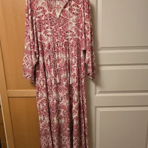 Vit och röd långklänning med mönster - Säljer en långklänning i vitt och rött med bohemiskt mönster. Klänningen har lång ärm, v-ringning och lös passform. Perfekt för dig som gillar en avslappnad och stilren look. Storlek M/L.