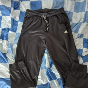 Svarta träningsbyxor från The North Face - Svarta träningsbyxor från The North Face med dragsko i midjan och logga på både framsidan och ena benet. Byxorna har meshfoder och är tillverkade i ett syntetmaterial som andas. Perfekta för träning eller chill. Passar s/m.