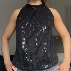 Svart glittrig ärmlös topp - Säljer en svart ärmlös topp med glittriga paljetter. Toppen har hög hals och ett lätt rynkat tyg framtill. Perfekt för dig som vill sticka ut med lite extra glitter.
