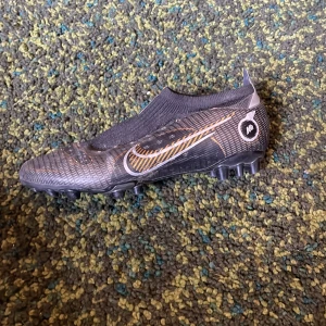 Svarta Nike fotbollsskor med gulddetaljer - Säljer ett par svarta Nike fotbollsskor med gulddetaljer och vit swoosh. Skorna har låg profil, plös utan snörning och mönstrad ovandel. Perfekta för spel på konstgräs eller gräsplan. Skorna är väl använda och dobbarna har fått lite slitage men inget jag har märkt unde användning. Nypris 2500 mitt pris 300