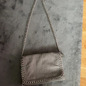 Tiamo axelväska  - Säljer en snygg grå axelväska från Tiamo med grov silverkedja som axelrem och runt kanterna. Väskan har dragkedja både på insidan och utsidan samt ett coolt, lite skrynkligt skinnliknande material. Perfekt för dig som gillar stilrena accessoarer med edge.