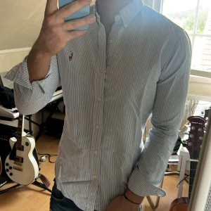 Blårandig skjorta från Ralph Lauren - Säljer en klassisk blårandig skjorta från Ralph Lauren med broderad logga på bröstet. Skjortan har lång ärm, knappar framtill och en snygg krage. Perfekt för dig som gillar stilrena och tidlösa plagg.