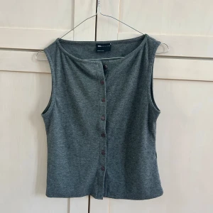 Grå ribbad linnetopp från ASOS DESIGN - Säljer en grå ribbad linnetopp från ASOS DESIGN med knappar framtill. Toppen är ärmlös och har en enkel, rak passform. Perfekt att matcha med jeans eller kjol för en avslappnad look. Aldrig använt, ej genomskinlig, sticks ej