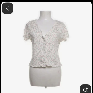Blommig ribbad topp från Monki - Supersöt kortärmad topp från Monki med blommigt mönster i rosa och grönt på vit botten. Toppen är ribbad, har v-ringning och knappar framtill. Perfekt för en fräsch och somrig look!