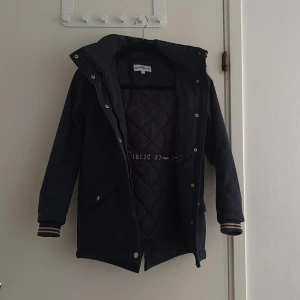 Mörkblå jacka - Snygg mörkblå parkajacka med avtagbar huva och fuskpäls. Jackan har quiltat innerfoder, guldiga knappar och dragkedja, samt ribbade muddar med randiga detaljer. Perfekt för kyliga dagar.