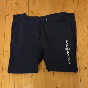 Marinblå mjukisbyxor från Sail Racing - Mörkblå mjukisbyxor från Sail Racing med vit logga längs ena benet. Byxorna har dragsko i midjan och bakficka. Perfekta för en avslappnad stil och riktigt sköna att ha på sig. Storlek L men sitter som M. 