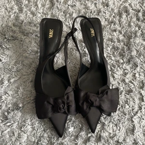 Svarta pumps med rosett från Zara - Säljer ett par eleganta svarta pumps från Zara med spetsig tå och stor rosett framtill. Skorna har öppen häl och smal klack, perfekta för att lyfta vilken outfit som helst.