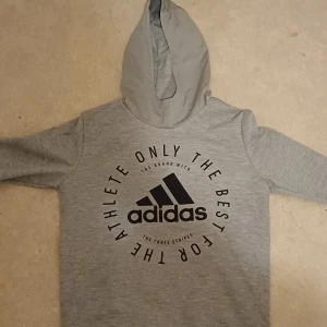 Grå hoodie från Adidas - Säljer en grå hoodie från Adidas med svart logga och text på bröstet. Tröjan har huva och långa ärmar, perfekt för sport eller chill. Mjuk och bekväm med klassisk passform. Adidas-tryck framtill ger en sportig vibe.