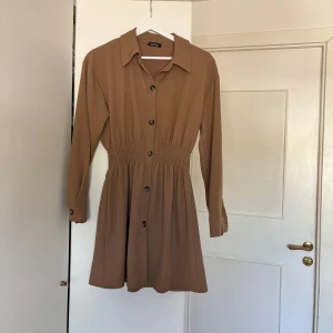 Brun skjortklänning med knappar - Säljer en snygg brun skjortklänning från BOOHOO med långa ärmar och knappar framtill. Klänningen har markerad midja med smock och klassisk krage. 