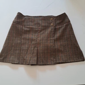 Brun rutig minikjol med shorts - Supersnygg brun och beige rutig minikjol från Hot Kiss med knappar framtill och inbyggda svarta shorts under. Midjemått är cirka 37 cm.