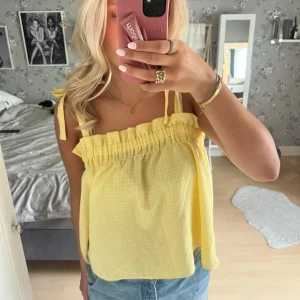 Gul linnetopp med rosettband - Söt gul linnetopp med smala axelband som knyts i rosetter. Toppen har en rynkad kant upptill och är luftig i modellen, perfekt för varma dagar.