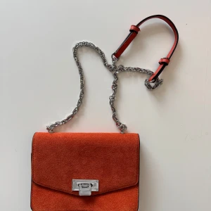 Orange axelväska i mocka med silverkedja - Säljer en snygg orange axelväska i mocka med silverfärgad kedjerem och metallspänne framtill. Väskan har en stilren, rektangulär form och är perfekt för att lyfta en outfit med lite färg. Aldrig använd 